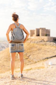 Vertical photo of a woman in summer clothes standing contemplating the landscape with a medieval castle in the background.