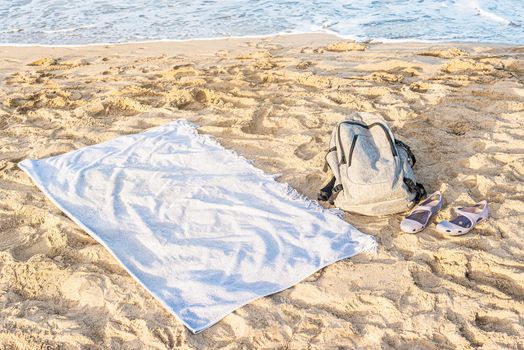 From above of white towel and backpack lying on sandy coast of waving sea in summer sunny day during vacation