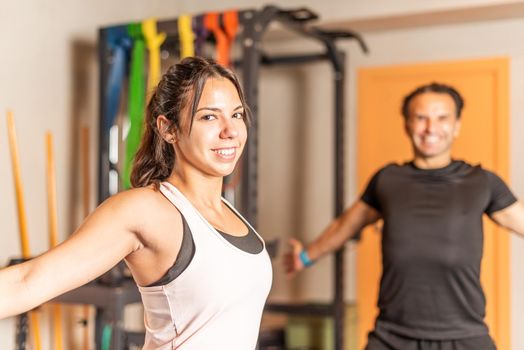 Close-up of a sportswoman stretching her arms back while looking at the camera with a sportsman behind her also looking at the camera. Concept of people in gym.