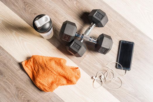 Top view of dumbbells, a bottle of water, a towel and a cell phone with headphones connected against a wooden background. Concept of equipment of gym and personal stuff.