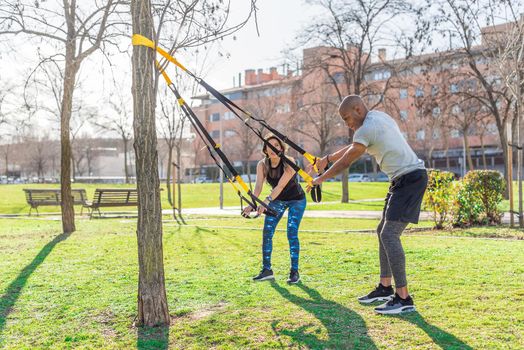 Fitness couple doing arms exercise with trx fitness straps in park. Multi-ethnic people exercising outdoors.