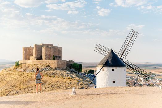 Young woman standing on a hill next to a windmill contemplating a castle on a sunny day in Consuegra, Spain.