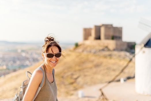 Young woman with sunglasses looking at the camera with a smile with an ancient castle on top of a hill in the background of the image out of focus in Toledo, Spain