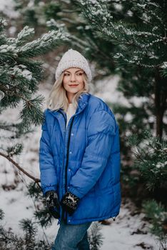 Young beautiful woman posing in forest during winter season. Attractive kind blonde girl smiling, lady in blue coat and white knitted hat. High quality