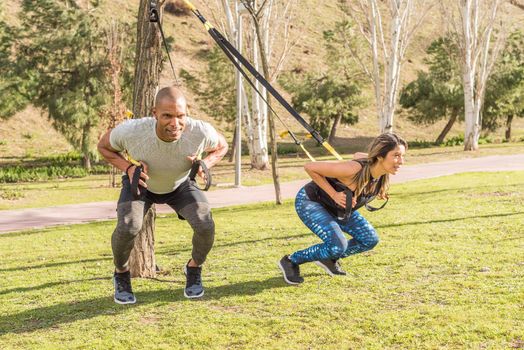 Athletic partners doing back exercise with trx fitness straps in park. Adult man and woman exercising outdoors.