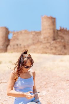 Vertical photo with selective focus on a young woman with sunglasses using mobile phone in front of a medieval castle. Almoncid castle in Spain