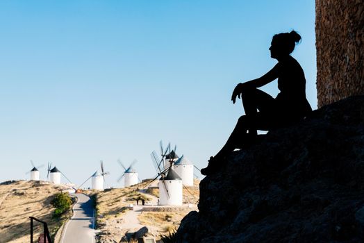 Silhouette of a woman sitting on the walls of a medieval castle viewing antique windmills in Toledo, Spain.