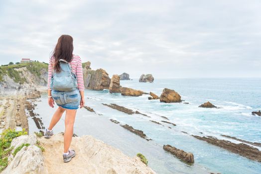 Back full body view of unrecognizable woman with backpack admiring seascape while standing on rocky cliff on sea coast during vacation