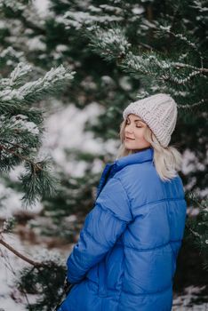 Young beautiful woman posing in forest during winter season. Attractive kind blonde girl smiling, lady in blue coat and white knitted hat. High quality
