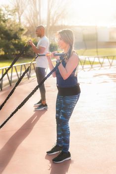 Standing atlhetic couple exercising arms with an elastic gym stick in the park. Multi-ethnic people exercising outside.