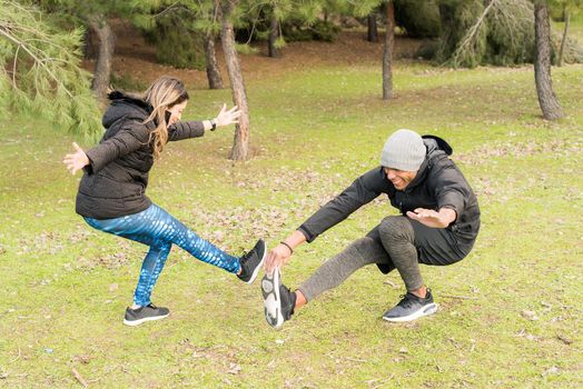 Fitness couple in warm clothes standing on one leg and stretching the other. Multi-ethnic couple outdoors.