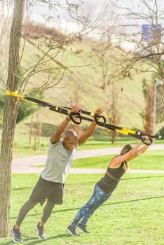 Full length view of fitness couple doing arm exercise with trx fitness straps in park. Multi-ethnic people exercising outdoors.