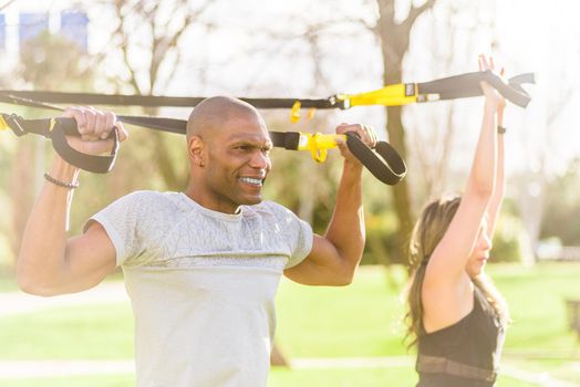 Fitness couple doing arms and biceps exercise with trx fitness straps in park. Multi-ethnic people exercising outdoors.