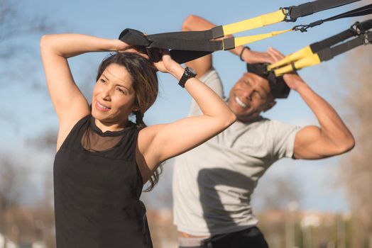 Athletic partners doing biceps and arms exercise with trx fitness straps in park. Multi-ethnic people exercising outdoors.