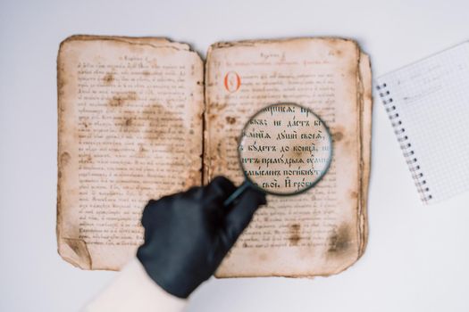 Historian scientist in gloves reading antique book with magnifying glass. Translation of religious literature. Manuscript with ancient writings. Treasures of the past. Museum piece. High quality