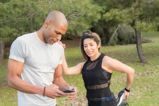 Fitness woman doing stretching leg on her partner who is using cell phone. Couple doing exercise outdoors together.