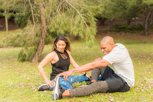 Sportsman stretching leg with her female partner in the park. Multi-ethnic people exercising outside.