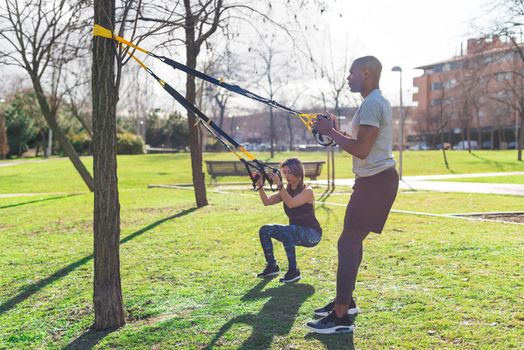 Couple of athletes exercising with trx fitness straps in the park. Multi-ethnic exercising outdoors.