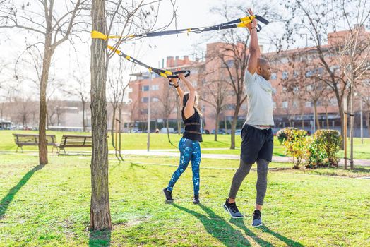 Fitness couple exercising with arms up and rotating the body with trx fitness straps in the park. Multi-ethnic people exercising outside.