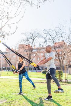 Fitness couple doing legs exercise with trx fitness straps in park. Multi-ethnic people exercising outdoors.