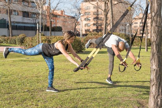 Athletic couple doing leg stretching exercise with trx fitness straps in park. Adult man and woman exercising outdoors.