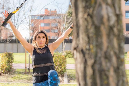 View near tree of a athlete woman exercising with trx fitness straps in the park. Adult woman exercising outside.