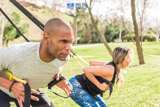 Close up view of an athletic partners doing back exercise with trx fitness straps in park. Adult man and woman exercising outdoors.