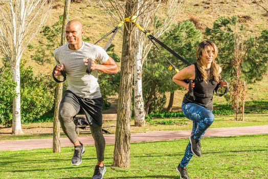 Fitness couple doing back legs exercise with trx fitness straps in park. Multi-ethnic people exercising outdoors.
