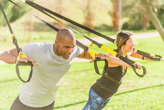 Fitness couple doing arm exercise with trx fitness straps in park. Multi-ethnic people exercising outdoors.