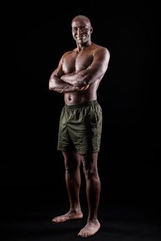 Adult African American in shorts standing with arms crossed. Muscular adult male with bare torso looking at camera on a black background.