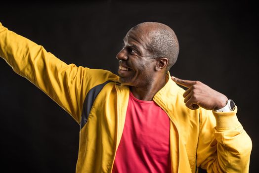 African American man standing looking and pointing to the side. Adult male in yellow jacket and red t-shirt in a studio with black background.