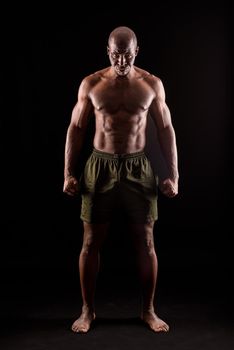 Muscular adult male standing looking at camera with serious expression with arms down and fists clenched. African American adult male standing dressed only in shorts on a black background.