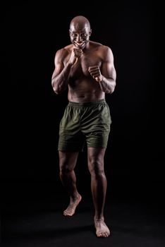 Muscular adult male smiling standing in defensive boxing pose. African American man wearing only shorts in a studio with black background.