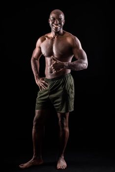 Cheerful adult muscular man standing looking at camera with a hand on waist and doing thumbs up with the other hand. African American adult male standing dressed only in shorts on a black background.