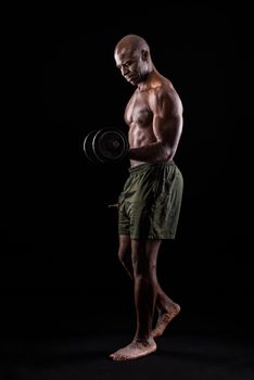 Muscular man standing doing exercise for biceps with dumbbell on a black background. African american adult in shorts doing exercise for arms.