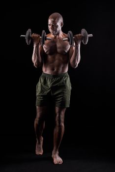 Front view of a muscular man standing doing exercise for biceps with dumbbells on a black background. African american adult in shorts doing exercise for arms.