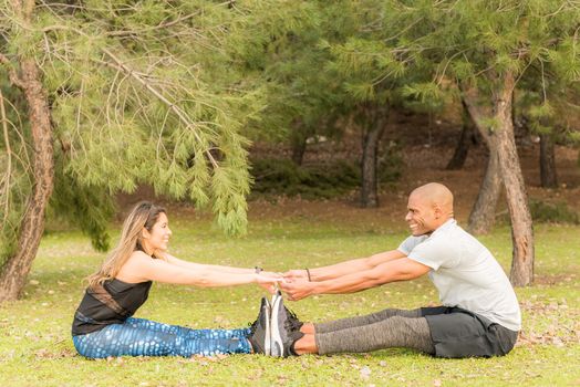 Sporty couple doing exercises together while sitting and holding hands. Multi-ethnic people exercising outside.