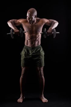 Front view of a muscular man standing doing triceps exercise with both arms with dumbbells on a black background. African american adult in shorts doing exercise for arms.