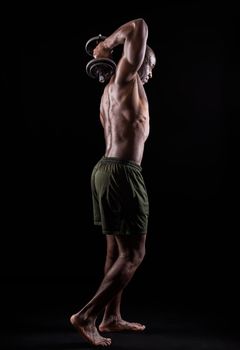 Side back view of a muscular man standing doing triceps exercise with dumbbells with arm raised on a black background. African american adult in shorts doing exercise for arms.