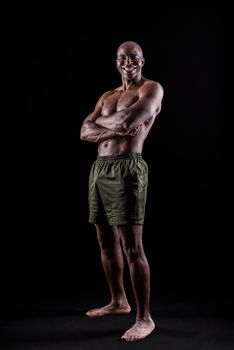 African American man standing in shorts with arms crossed in a black background. Muscular adult male with bare torso looking at camera.