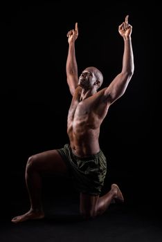 African American adult in shorts kneeling with arms extended upward. Adult male on black background with naked torso rolled up and striking a triumphant pose pointing upward.