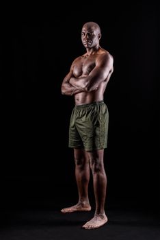 Adult African American in shorts standing with arms crossed looking at camera seriously on a black background. Muscular adult male with bare torso looking at camera on a black background.