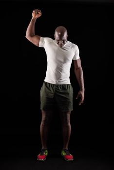 African American male standing with one arm raised and fist clenched while looking down. Muscular adult man in sportswear in a studio with black background.