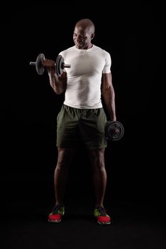Muscular man standing doing exercise for biceps with dumbbell on a black background. African american adult wearing t-shirt, shorts doing exercise for arms.