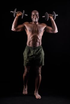 Athletic adult man doing shoulder exercise with dumbbells. Strong african american bodybuilder looking at camera and doing shoulder exercises on a black background.