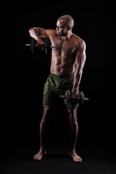 Front view of a muscular man standing doing triceps exercise with dumbbells on a black background. African american adult in shorts doing exercise for arms.