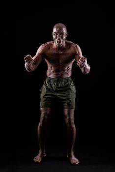 African American man facing front with fists clenched and shouting at camera. Athletic adult man standing dressed only in shorts with a black background.