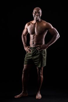 Cheerful adult muscular man standing looking at camera with hands on waist. African American adult male standing dressed only in shorts on a black background.