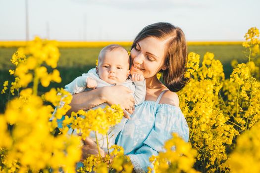 Young mother holding baby boy. Son having fun, smiling in yellow canola field. Love, family, joy concept.