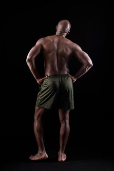 Back view of a muscular man standing with his hands on his waist. Bald adult African American man seen from behind on a black background in studio.